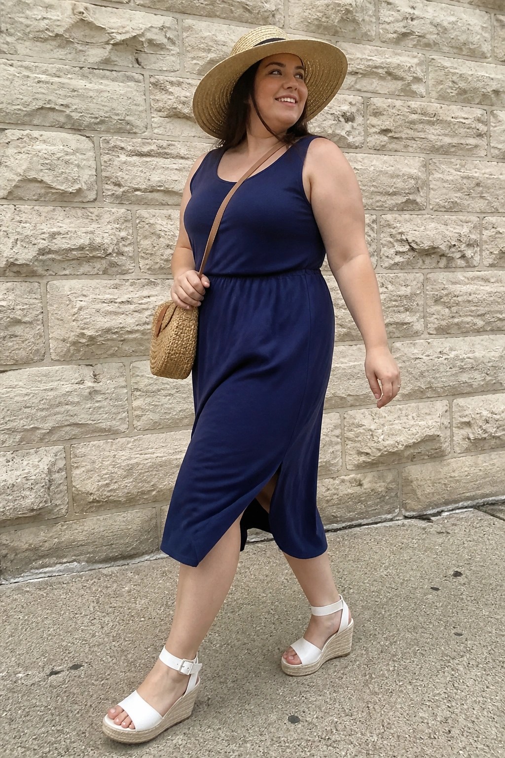 Plus size woman wearing a navy blue sleeveless midi dress with side slit, white wedge espadrille sandals, straw hat, and small woven crossbody bag standing against a stone wall.