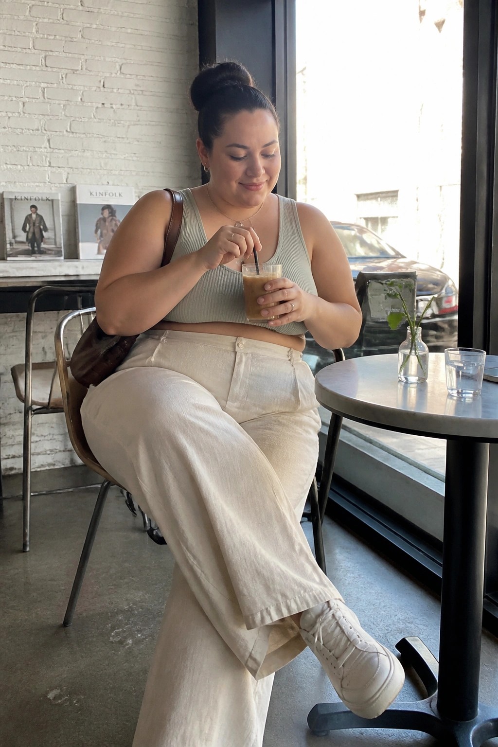 Woman wearing a sage green ribbed crop tank top with cream wide-leg linen pants, white sneakers, and a brown leather bag while seated at a cafe table holding an iced coffee.