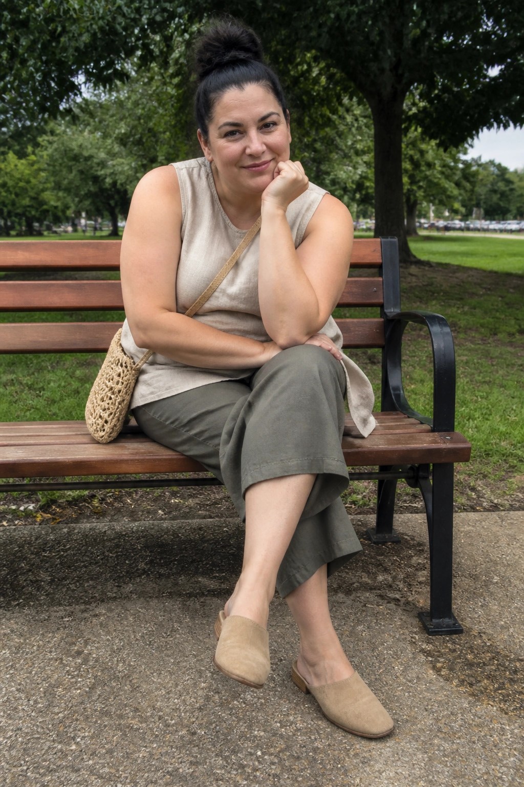 Woman seated on a park bench wearing a beige sleeveless linen top with side tie, olive green wide leg cropped pants, beige suede flat mules, and a woven crossbody bag.