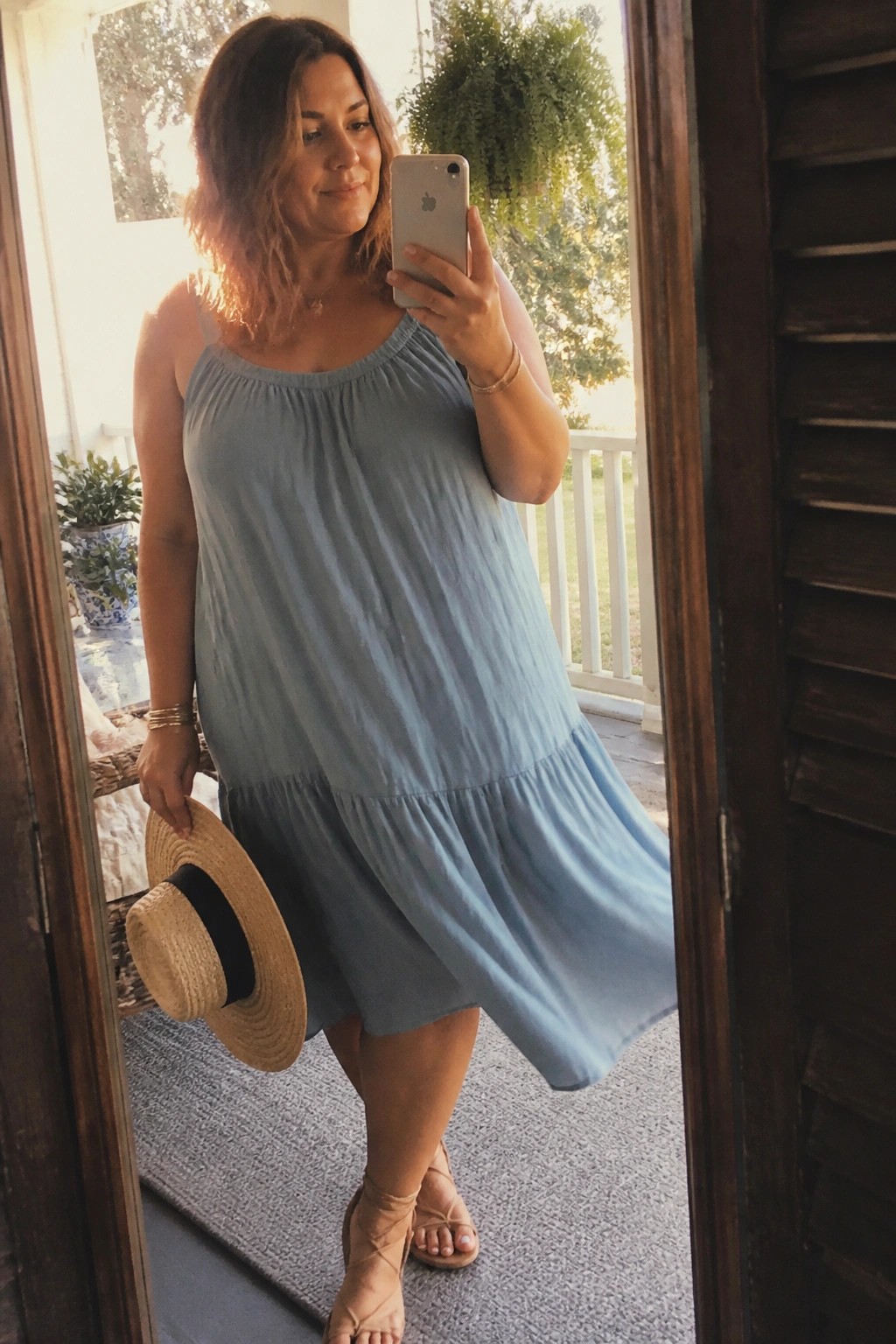 Woman wearing a light blue tiered midi sundress with thin straps and strappy beige sandals while holding a straw hat and phone in a doorway
