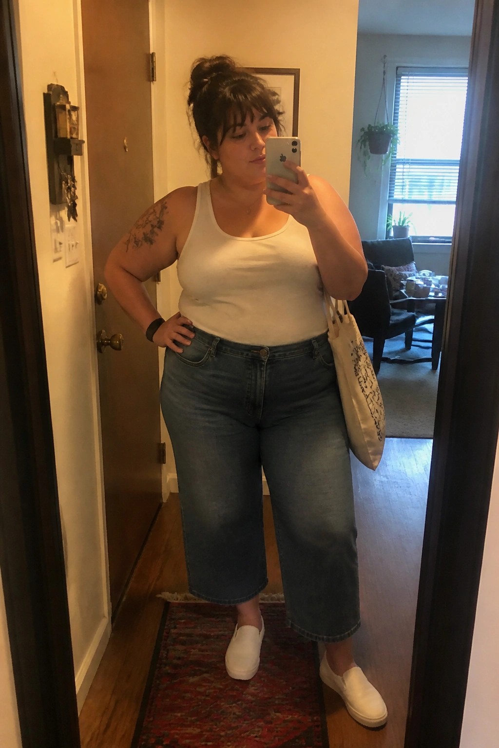 Plus size woman in mirror selfie wearing white ribbed tank top, blue wide leg cropped jeans, white slip on sneakers and carrying a beige tote bag.