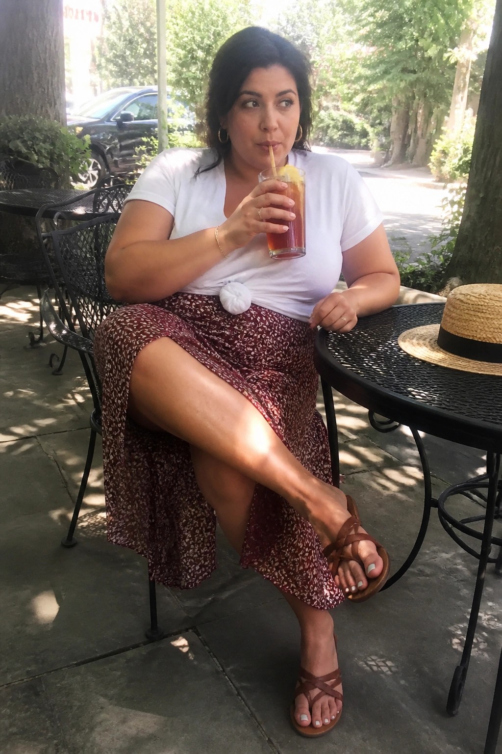 Plus size woman in a white short sleeve tee tied at the waist with a white pom pom accent and a burgundy floral print midi skirt paired with brown strappy sandals.