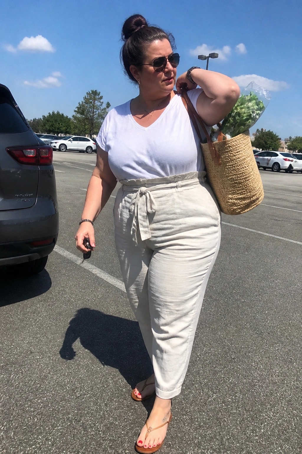 Woman in white tee and beige pants carrying woven straw bag in sunny lot