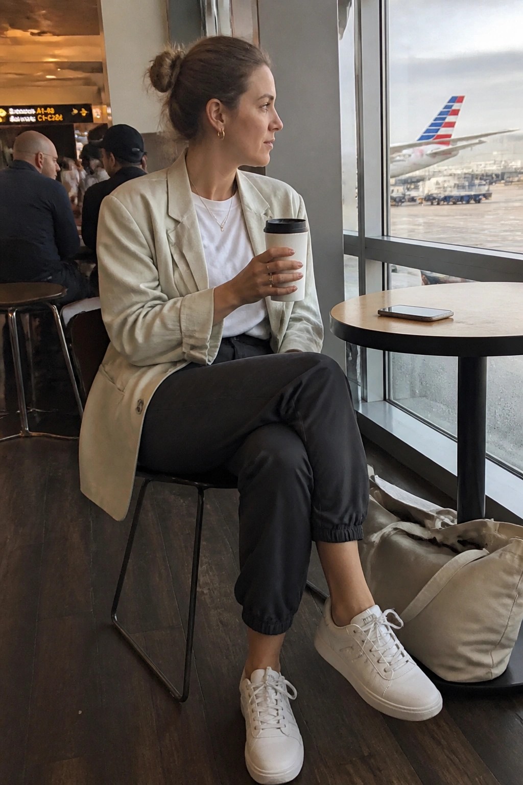 Woman wearing an oversized beige blazer over a white t-shirt with black cuffed jogger pants and white sneakers while holding a coffee cup in an airport setting.