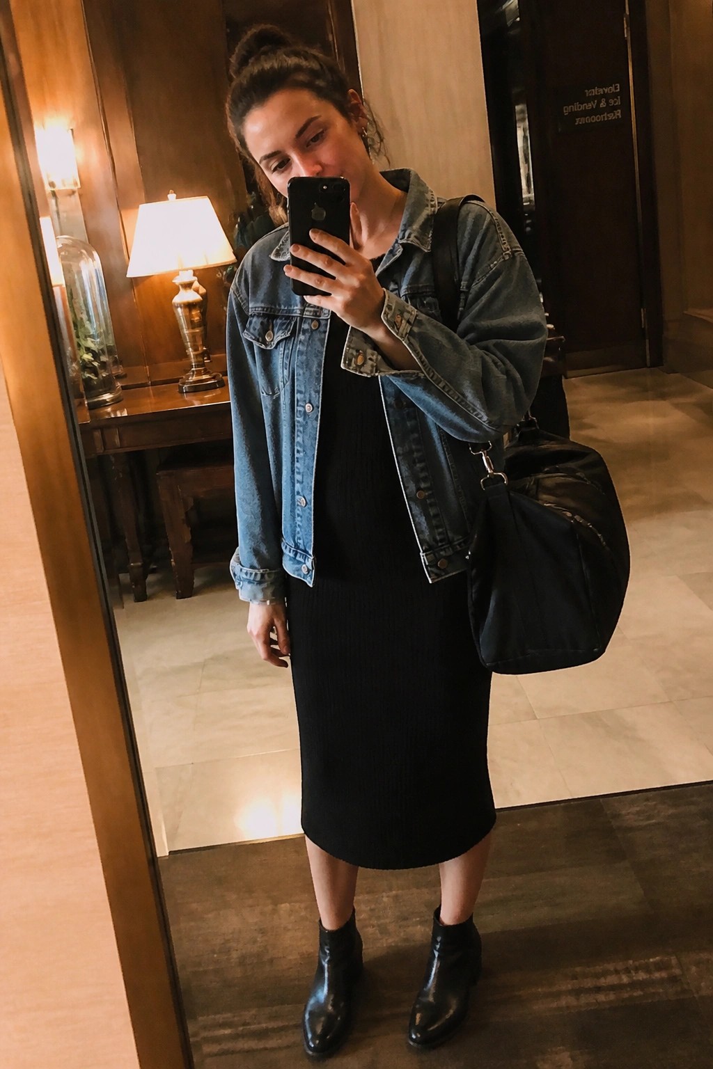 Woman in black ribbed midi dress layered under oversized blue denim jacket paired with black ankle boots and carrying a black bag while taking a mirror selfie.