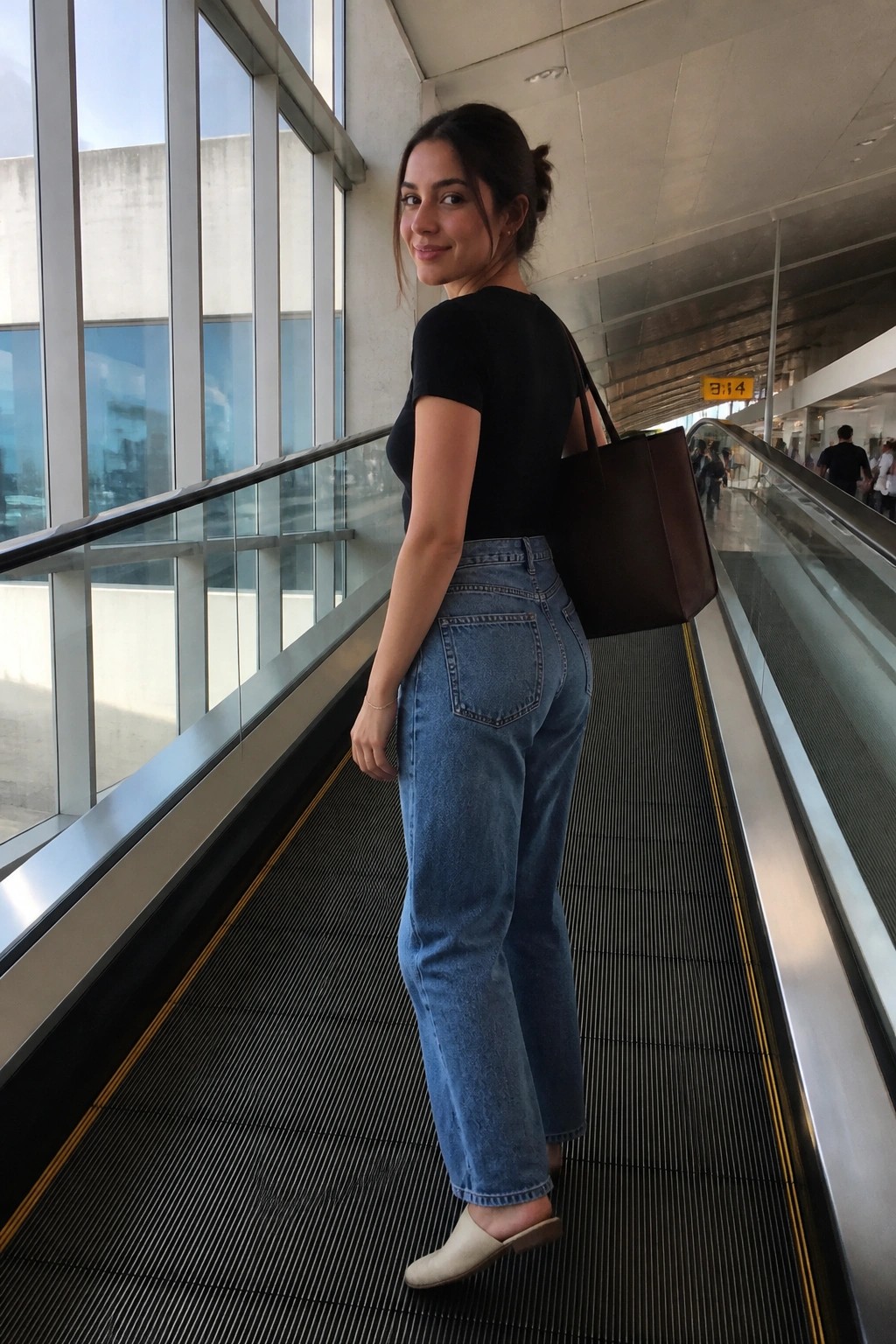 Woman in black top and jeans with brown bag on airport escalator looking back