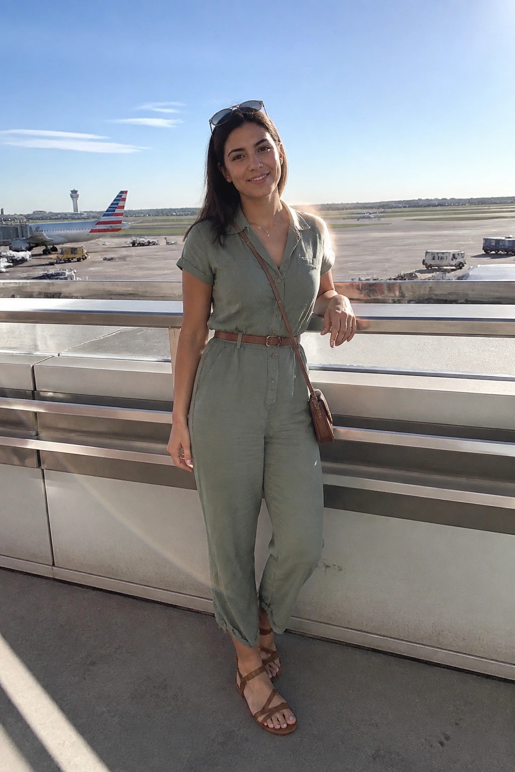 Woman in sage green jumpsuit at sunny airport overlooking runway and plane