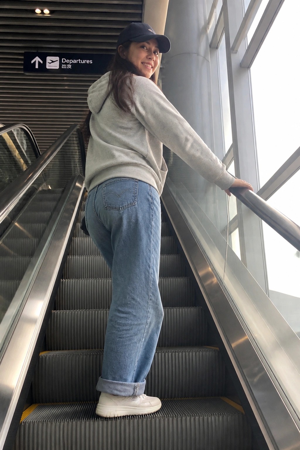 Woman in gray hoodie and jeans on airport escalator near departures sign