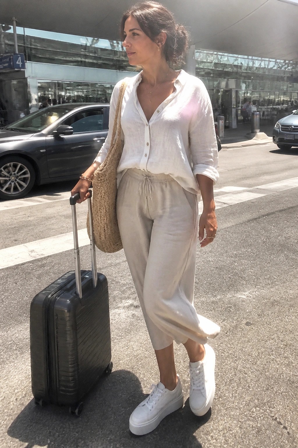 Woman in white linen button-up shirt tucked into beige cropped wide-leg pants with white platform sneakers, holding a black rolling suitcase and carrying a beige woven shoulder bag.