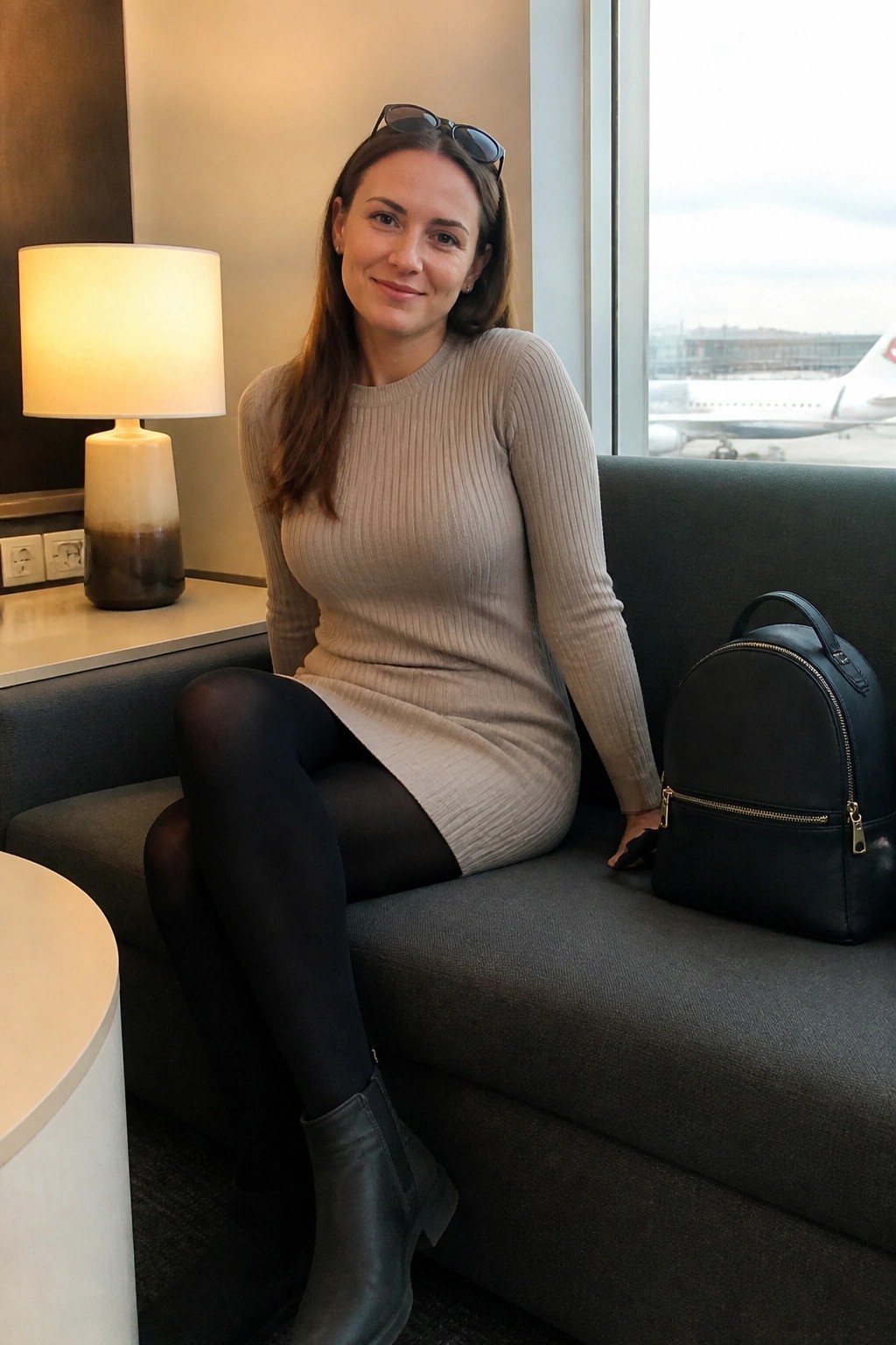 Woman in beige sweater dress seated on gray couch with black backpack near airport window