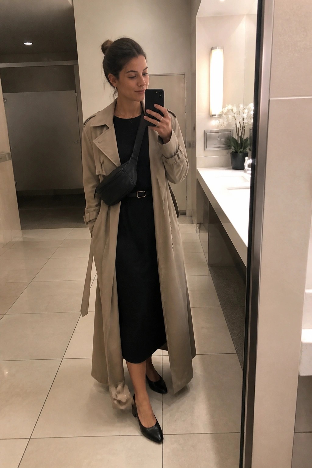 Woman wearing an open beige trench coat over a black midi dress paired with a black crossbody bag, slim belt, and pointed black flats while holding a phone for a mirror selfie.