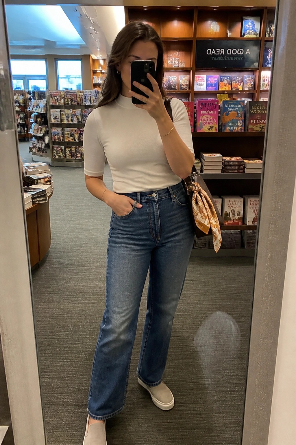 Woman in white top and blue jeans taking mirror selfie in bookstore