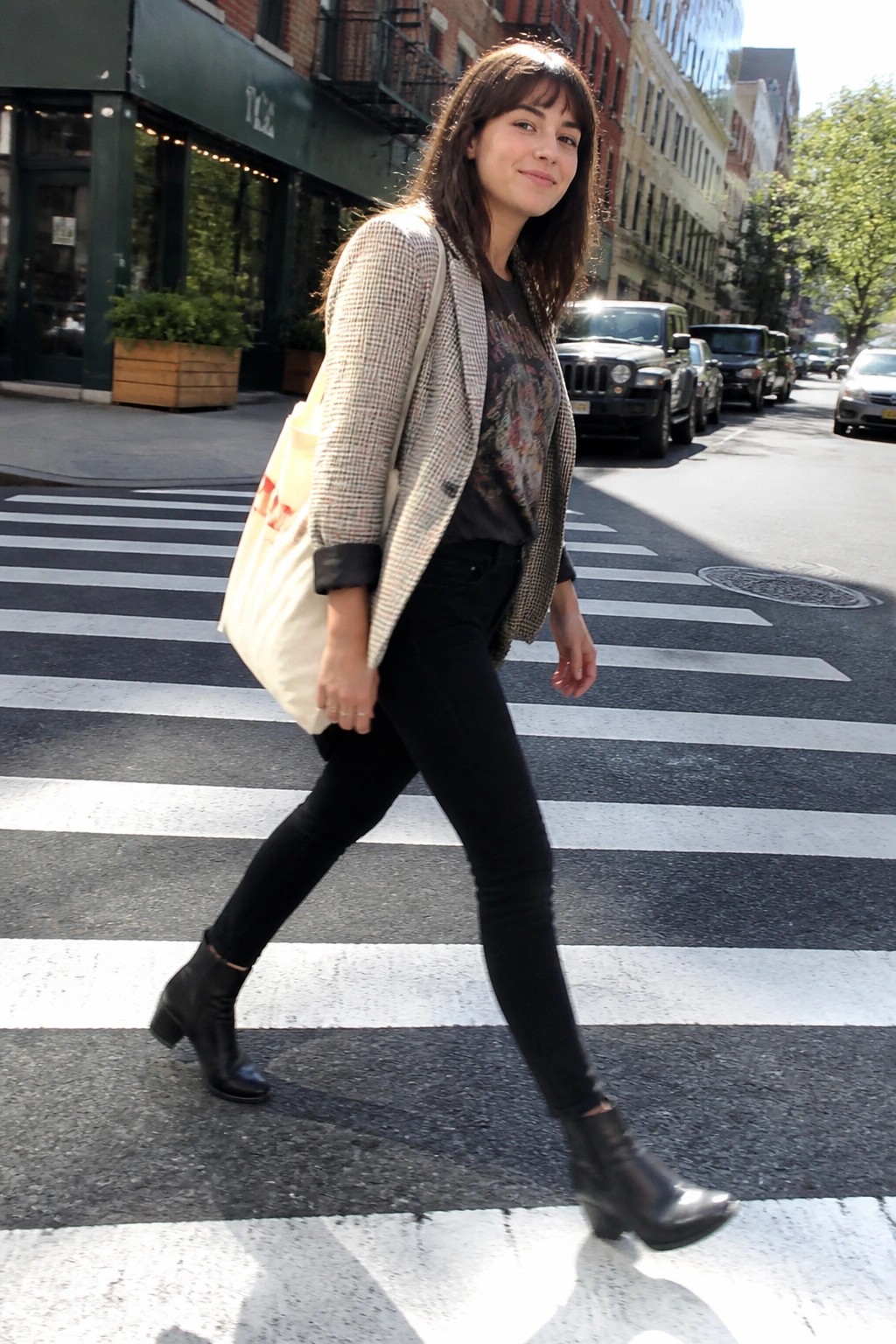 Woman in houndstooth blazer crosses sunny city crosswalk carrying tote bag