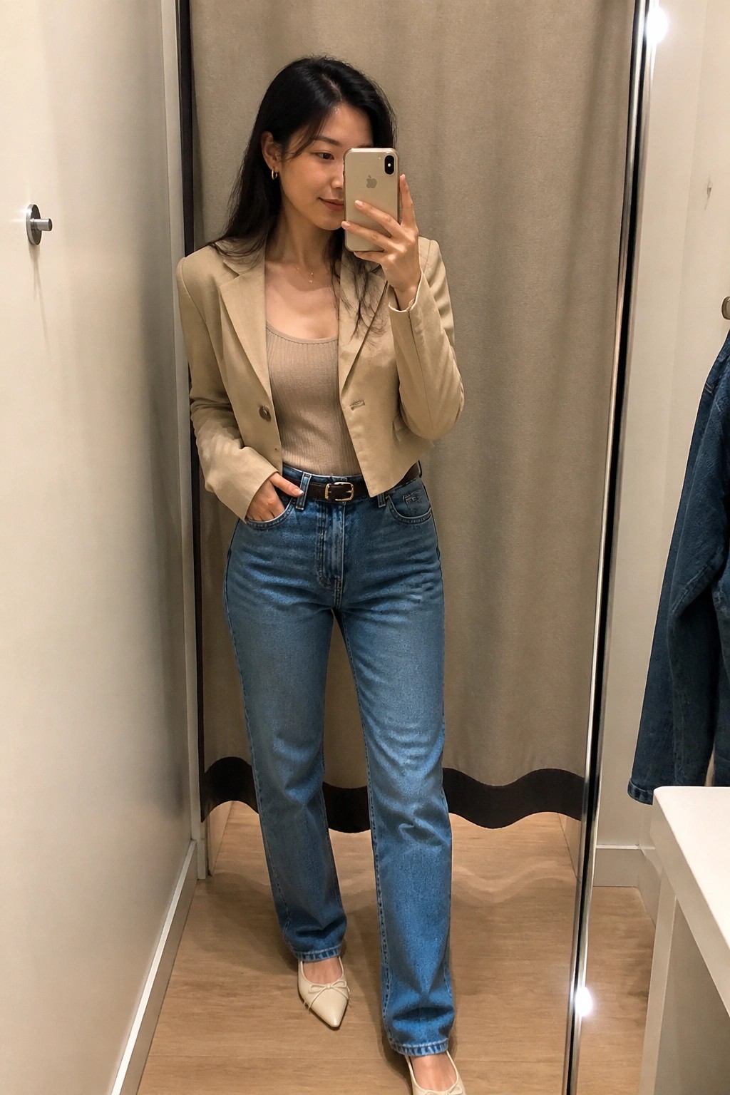 Woman in beige blazer and blue jeans taking mirror selfie in fitting room