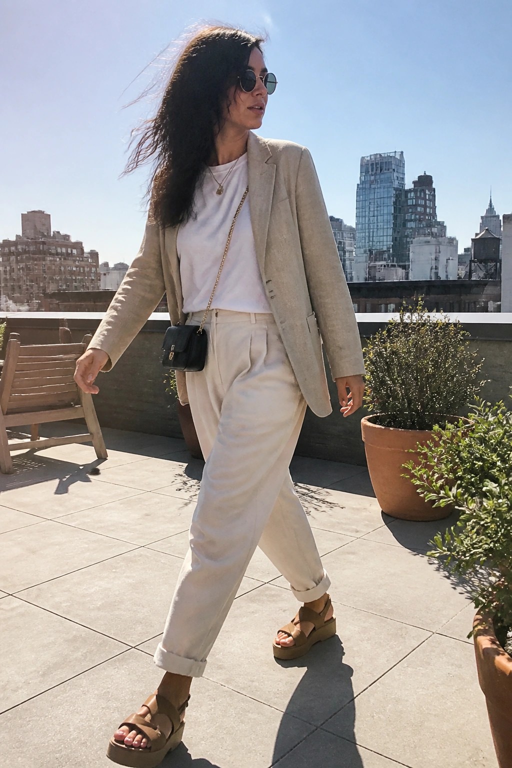 Woman in a beige blazer over a white tee with white pleated trousers cuffed at the ankles and brown platform sandals carrying a small black crossbody bag on a gold chain.