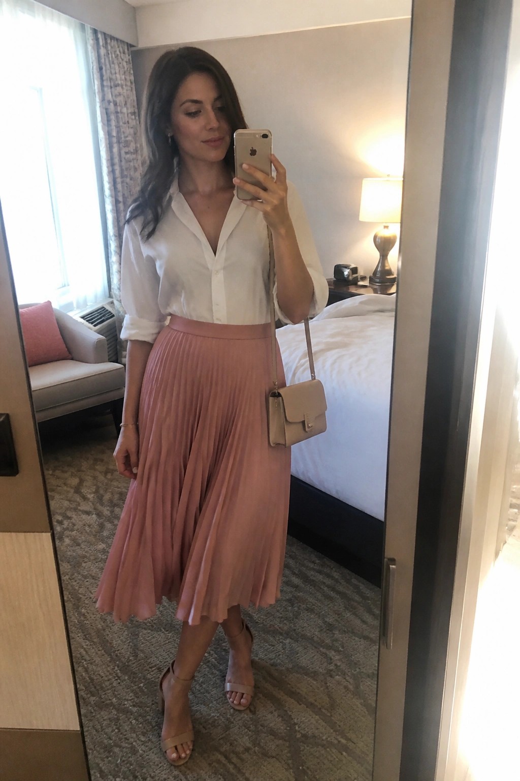 Woman in white blouse and pink pleated skirt taking mirror selfie in hotel room