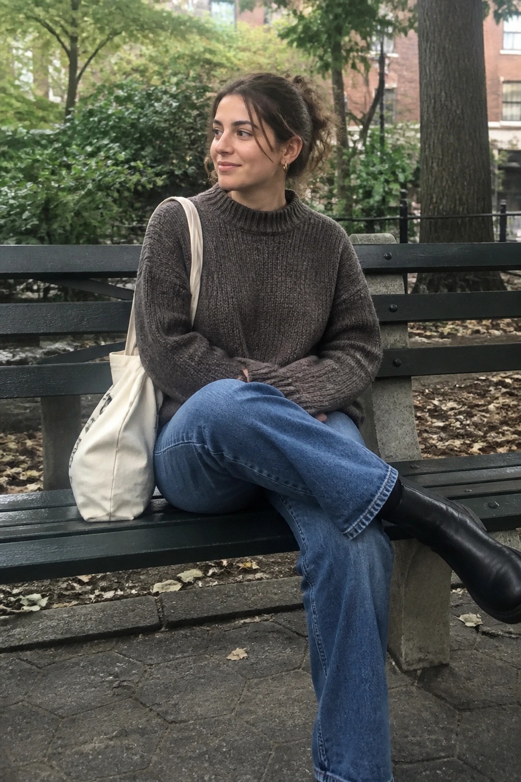 Woman in brown sweater and blue jeans with tote bag on park bench
