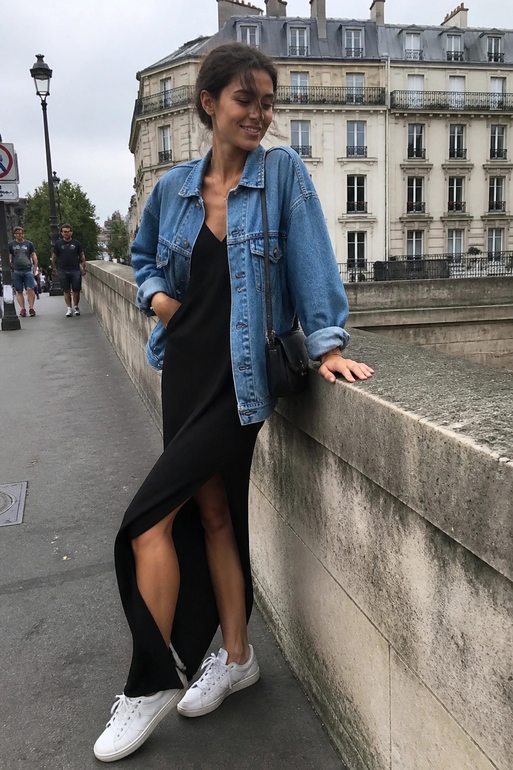 Woman in denim jacket over black dress leaning on stone bridge railing