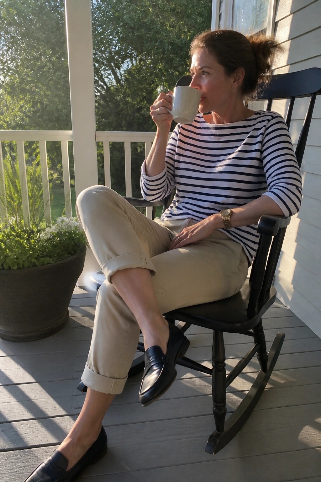 Woman in striped shirt sips from mug on black rocking chair on sunny porch