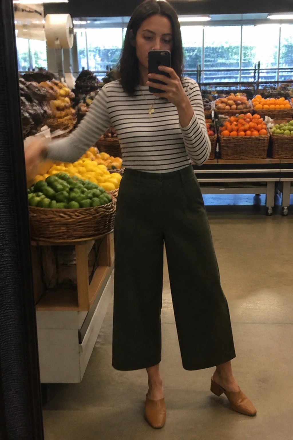 Woman in striped shirt and wide green pants taking mirror selfie in market.