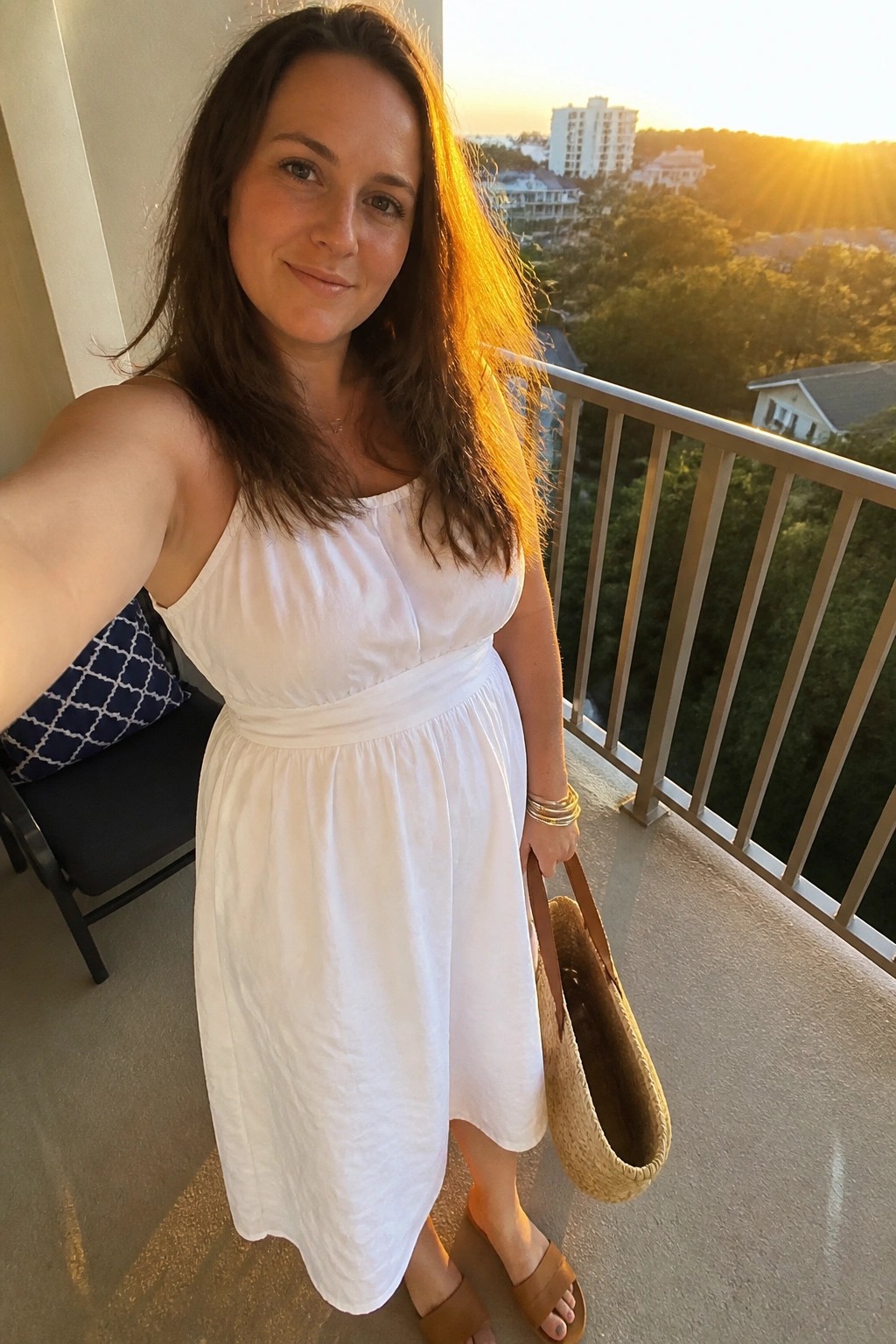 Woman wearing a white sleeveless midi dress with a smocked bodice and flowing skirt paired with brown slide sandals and carrying a woven straw tote bag.