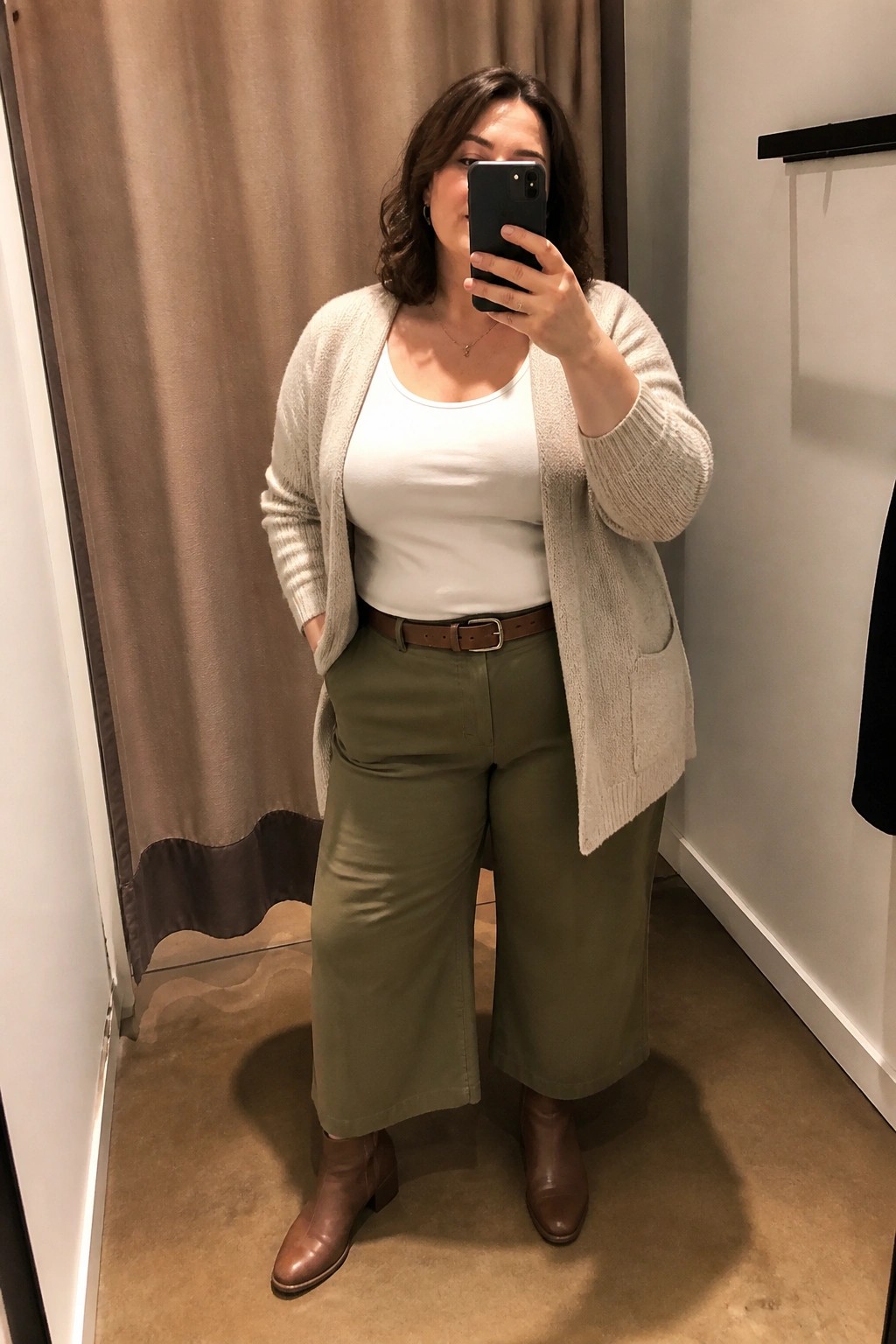 Plus size woman wearing a beige open cardigan over a white tank top with olive wide leg pants, brown belt, and brown ankle boots in a casual mirror selfie.