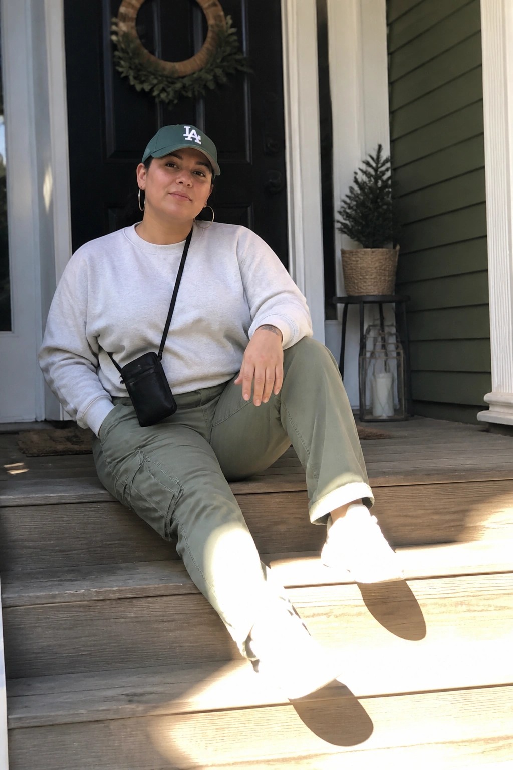 Woman in green LA cap, gray sweatshirt, olive pants on porch steps