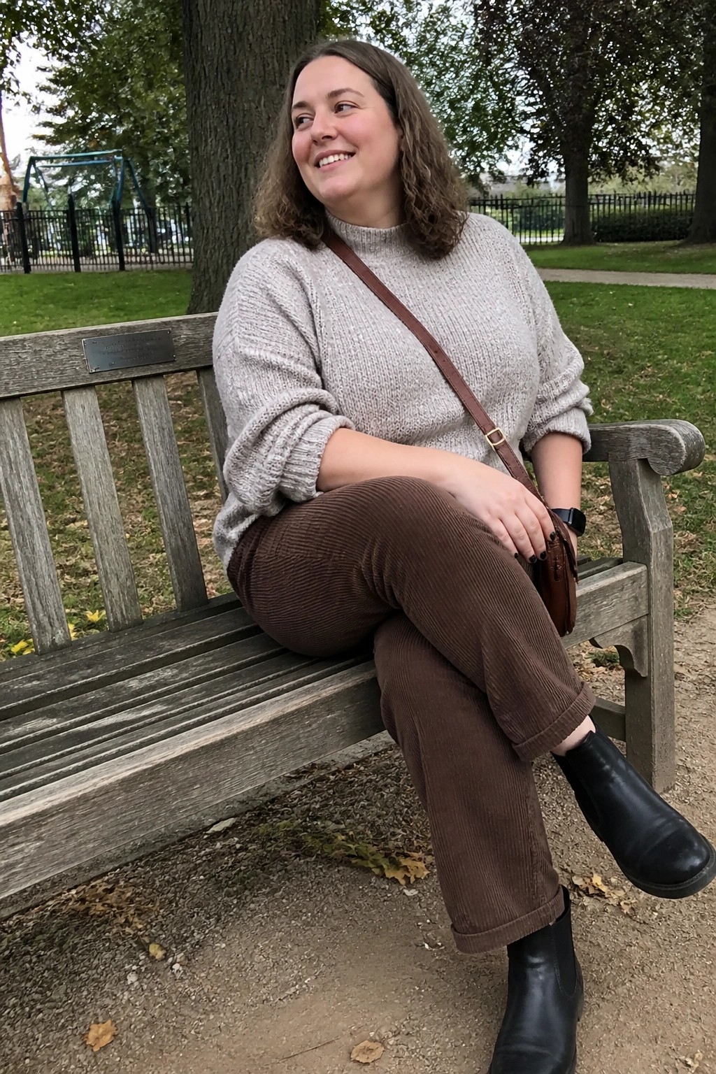 A plus size woman wearing a light gray chunky knit sweater, brown corduroy pants, and black ankle boots sitting on a wooden bench with a brown crossbody bag.