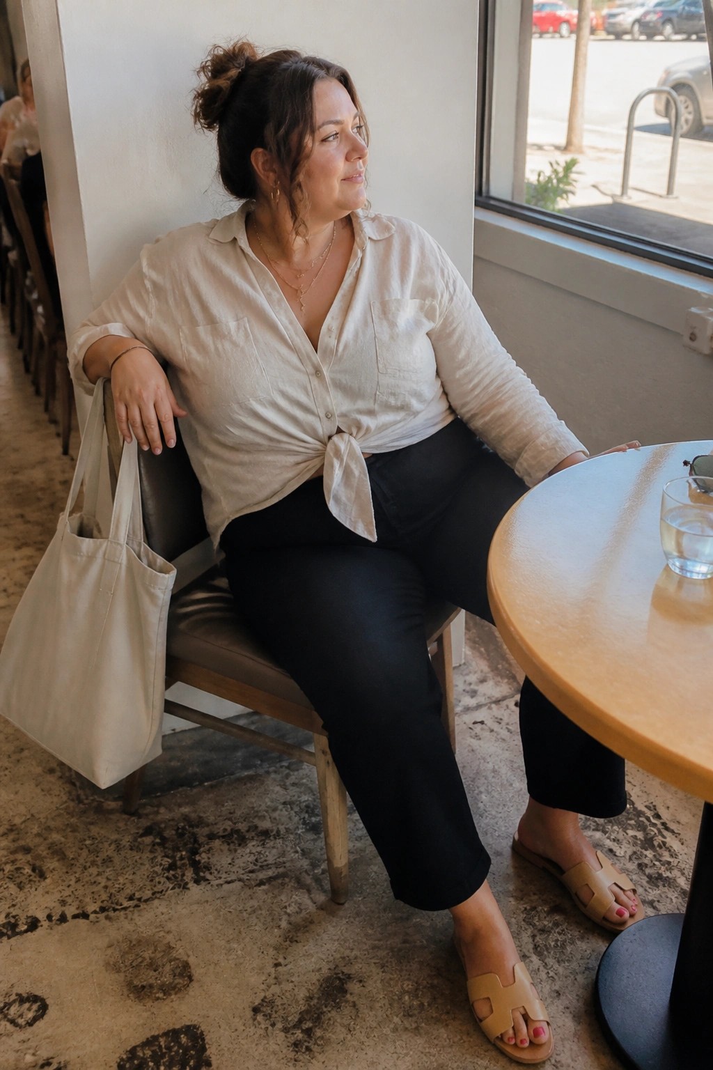 Woman in beige knotted shirt and black pants seated at cafe table with tote bag
