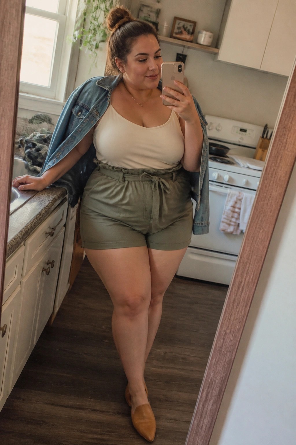 Plus size woman in olive green tie-waist shorts, cream tank top, and open denim jacket over her shoulders, wearing tan pointed flats while taking a mirror selfie in a kitchen.