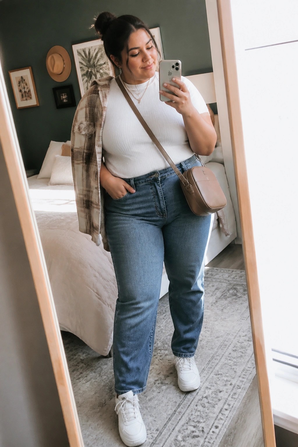 Woman in white ribbed top layered with open brown plaid shirt, blue jeans, white sneakers, and brown crossbody bag standing in a bedroom mirror selfie.