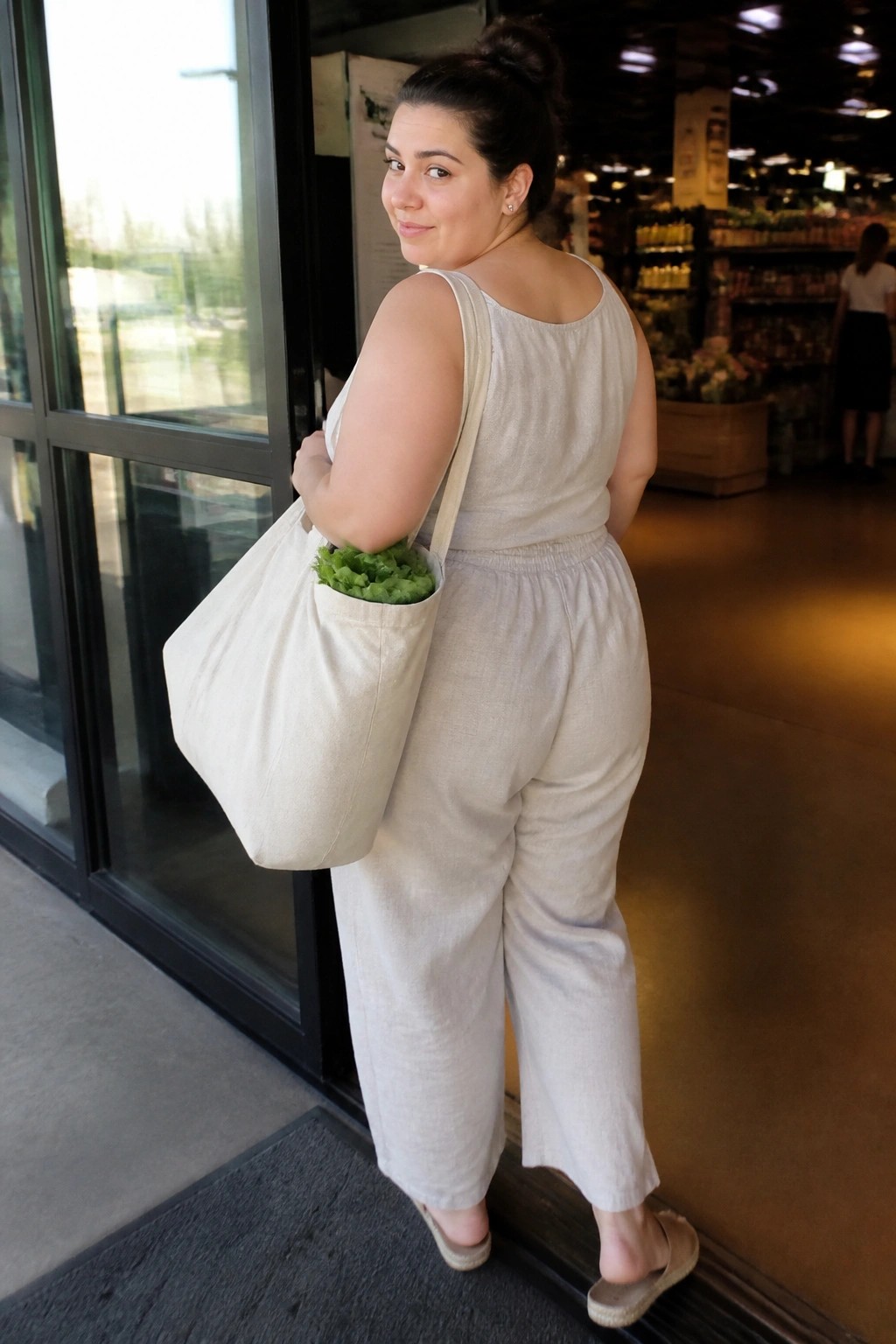 Woman in beige linen jumpsuit carries tote bag of greens at store entrance.