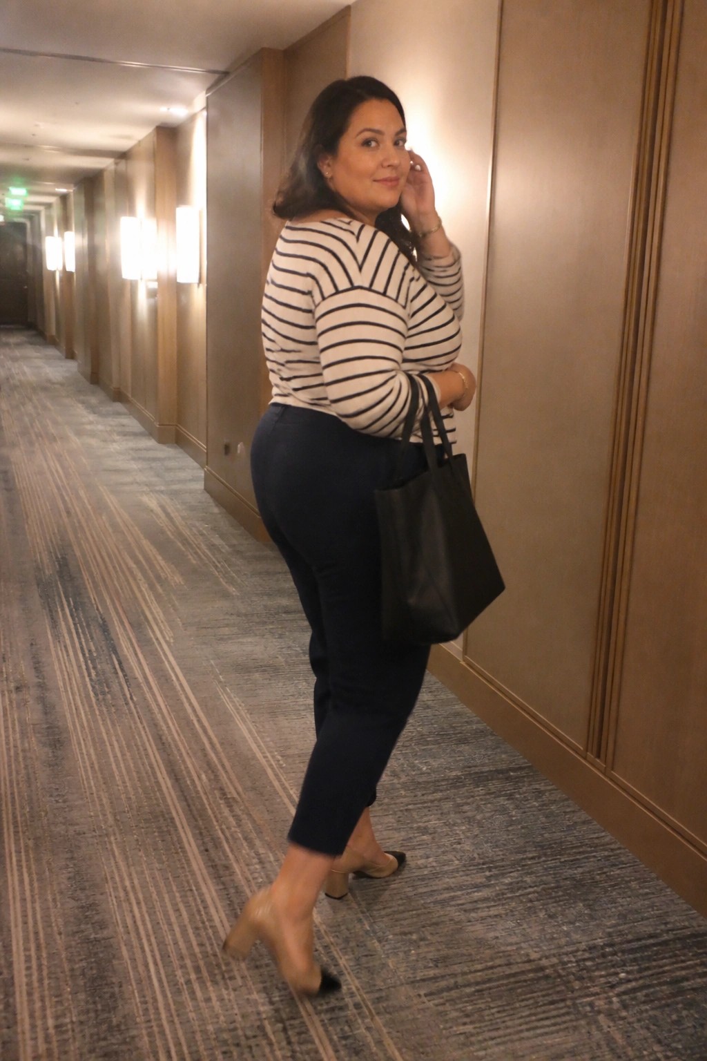 Woman in striped top and navy pants holds black tote in hallway