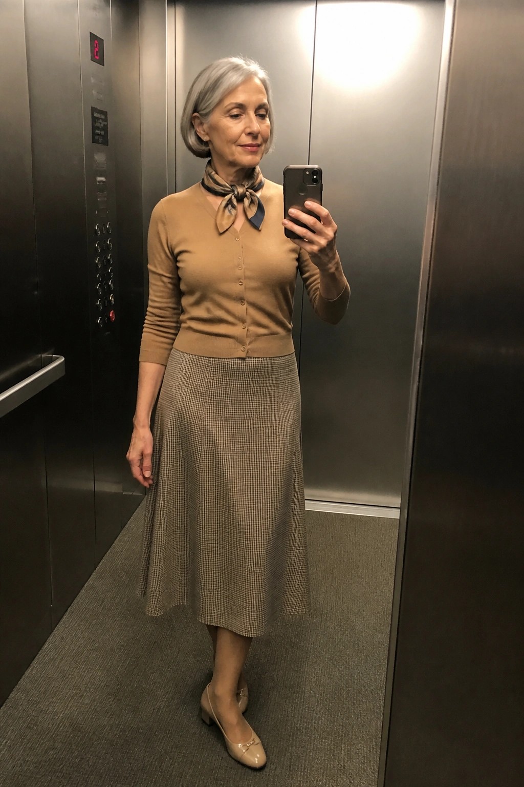 Gray-haired woman in tan cardigan and houndstooth skirt taking elevator selfie