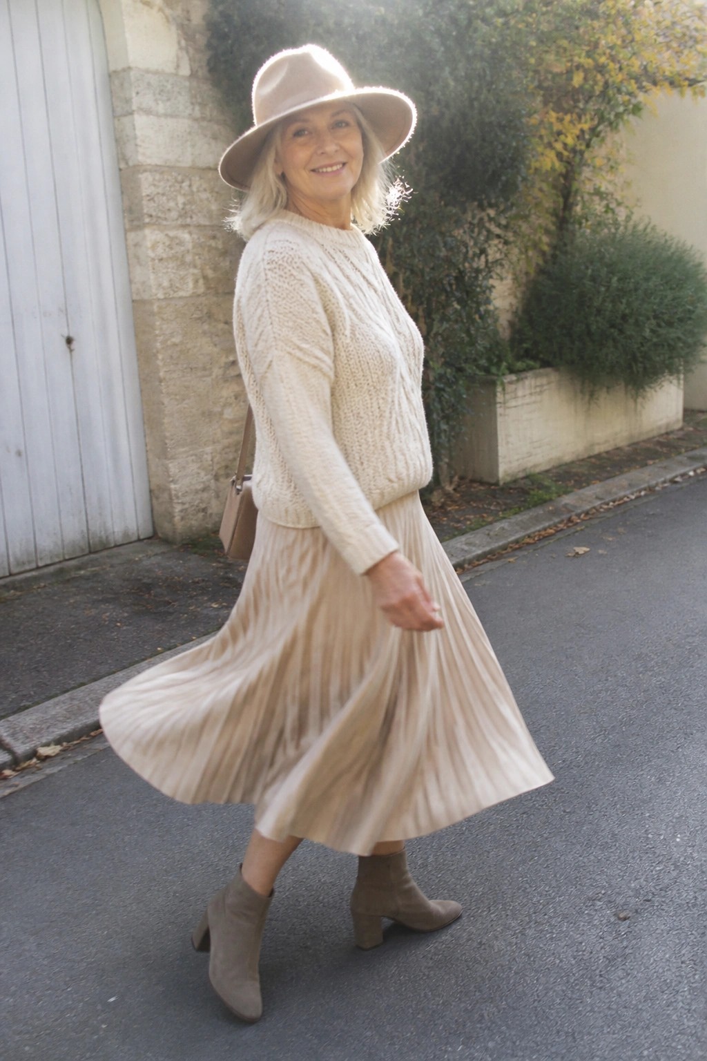 Smiling woman in wide hat, cream sweater, and flowing pleated skirt outdoors