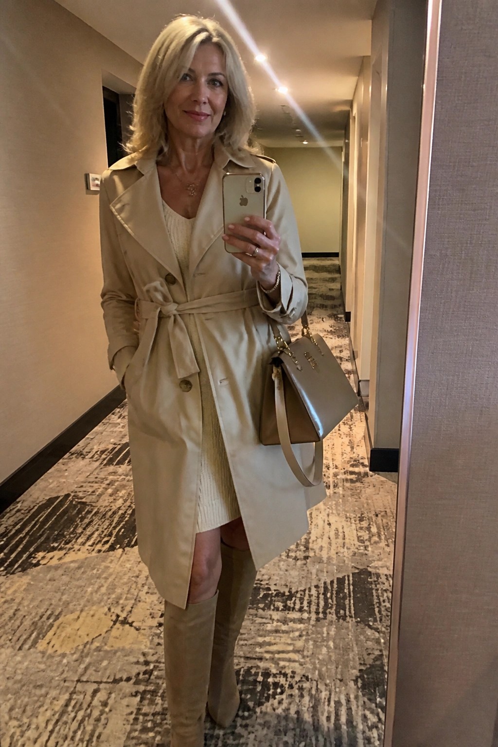 Blonde woman in beige trench coat and boots taking mirror selfie in hallway.