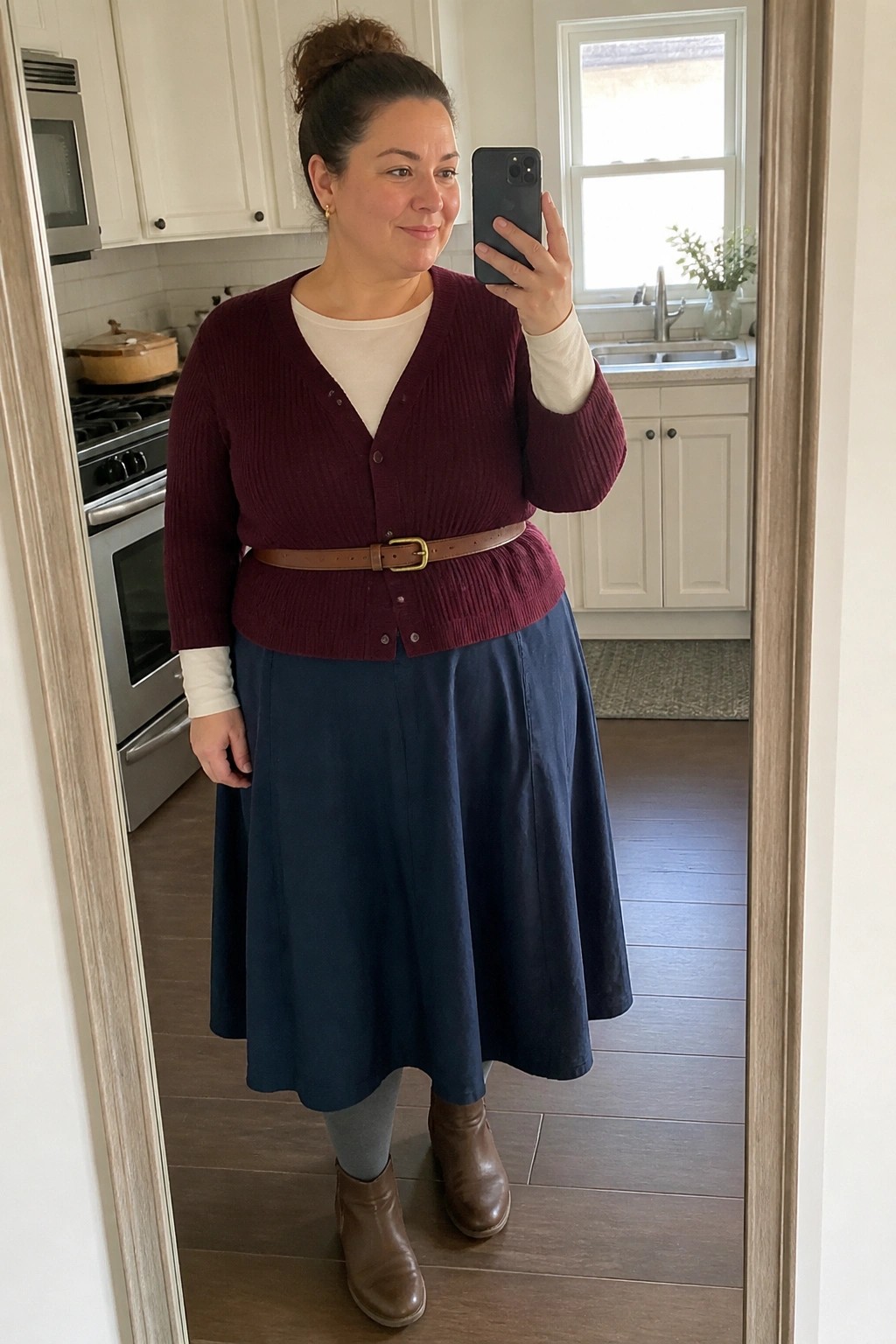 Woman in maroon cardigan and blue skirt taking mirror selfie in kitchen