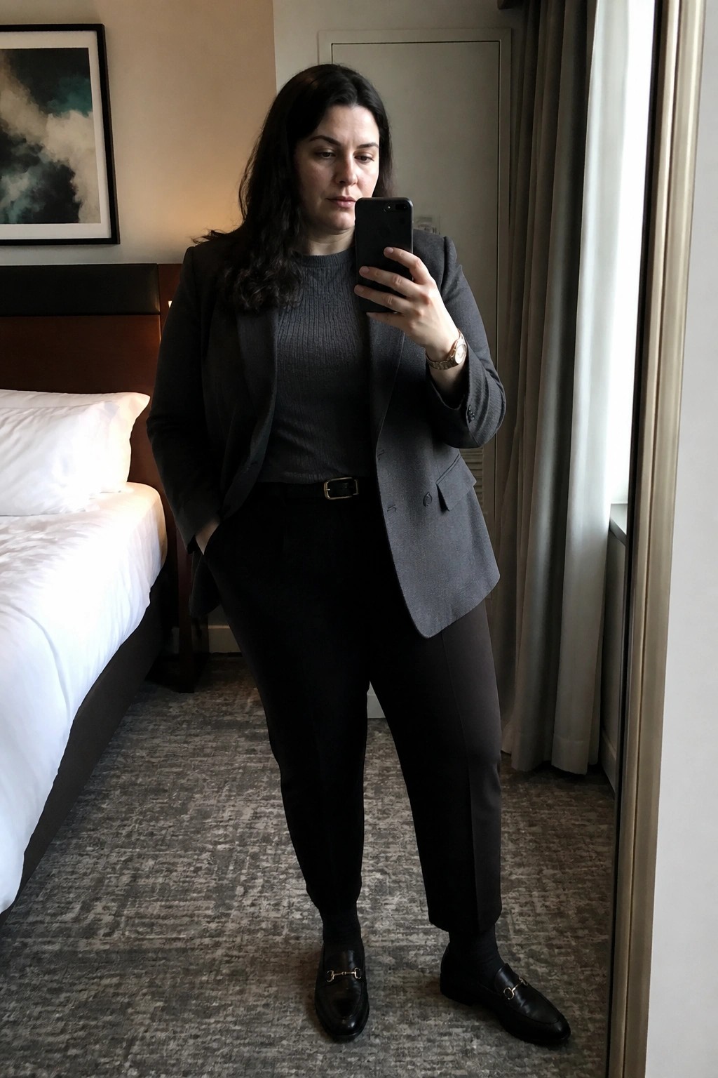 Woman in gray blazer and black pants taking mirror selfie in hotel room