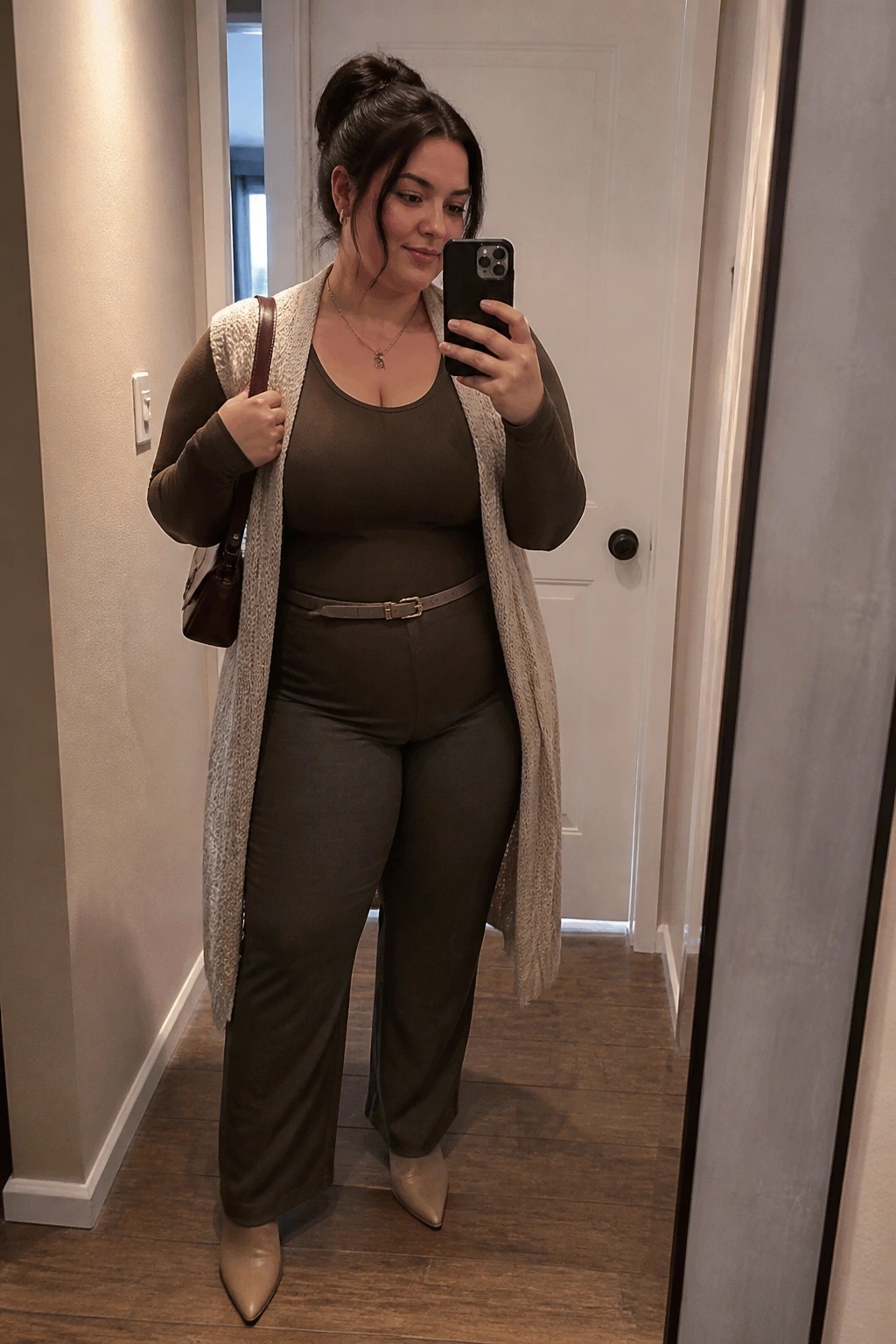 Woman wearing matching brown long sleeve top and wide leg pants layered with an open long beige knit cardigan belted at the waist paired with beige pointed toe boots and holding a phone for a mirror selfie.