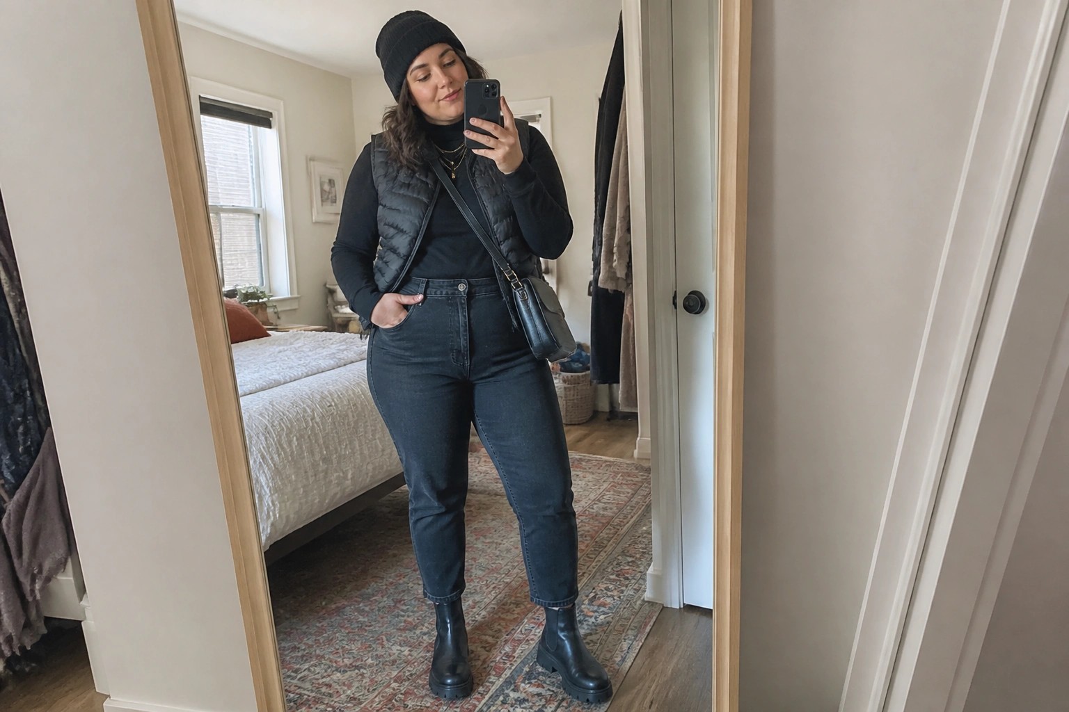 Woman in cream beanie, black vest, and jeans taking bedroom mirror selfie