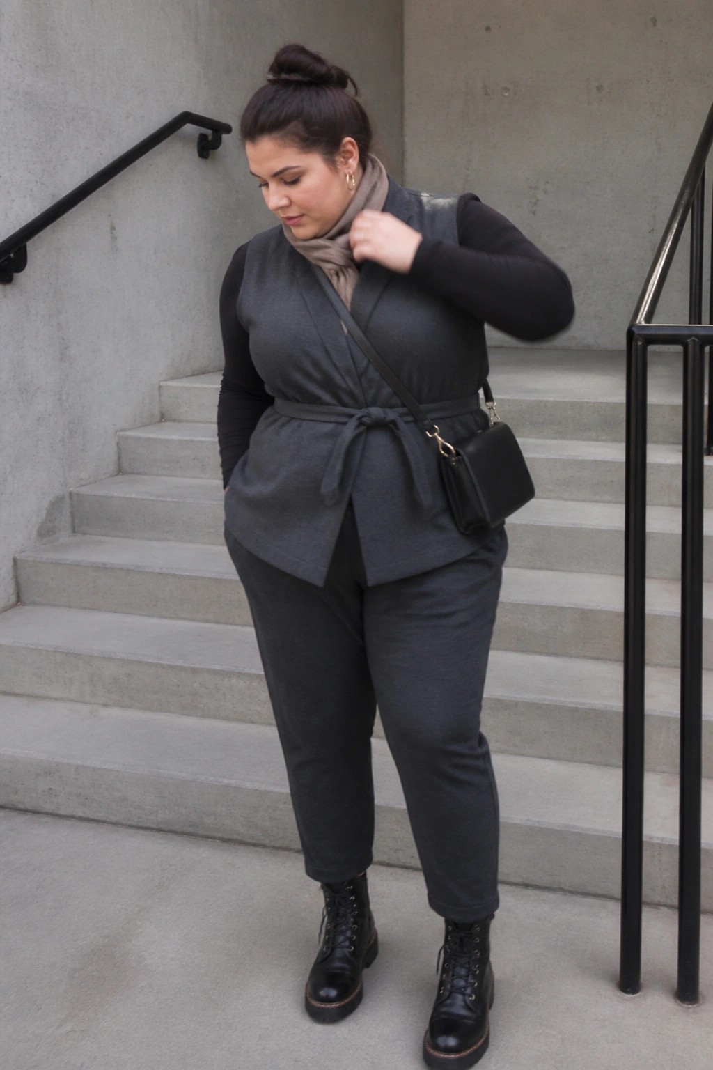 Plus size woman in dark gray wrap vest tied at waist over black long sleeve top paired with matching gray pants black combat boots black crossbody bag and light gray scarf.