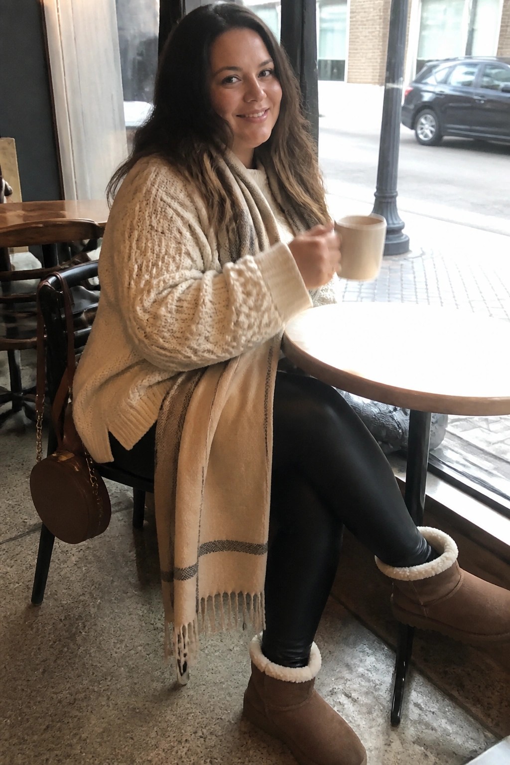 Woman in cream knit sweater and scarf holding mug at cafe table in Ugg boots