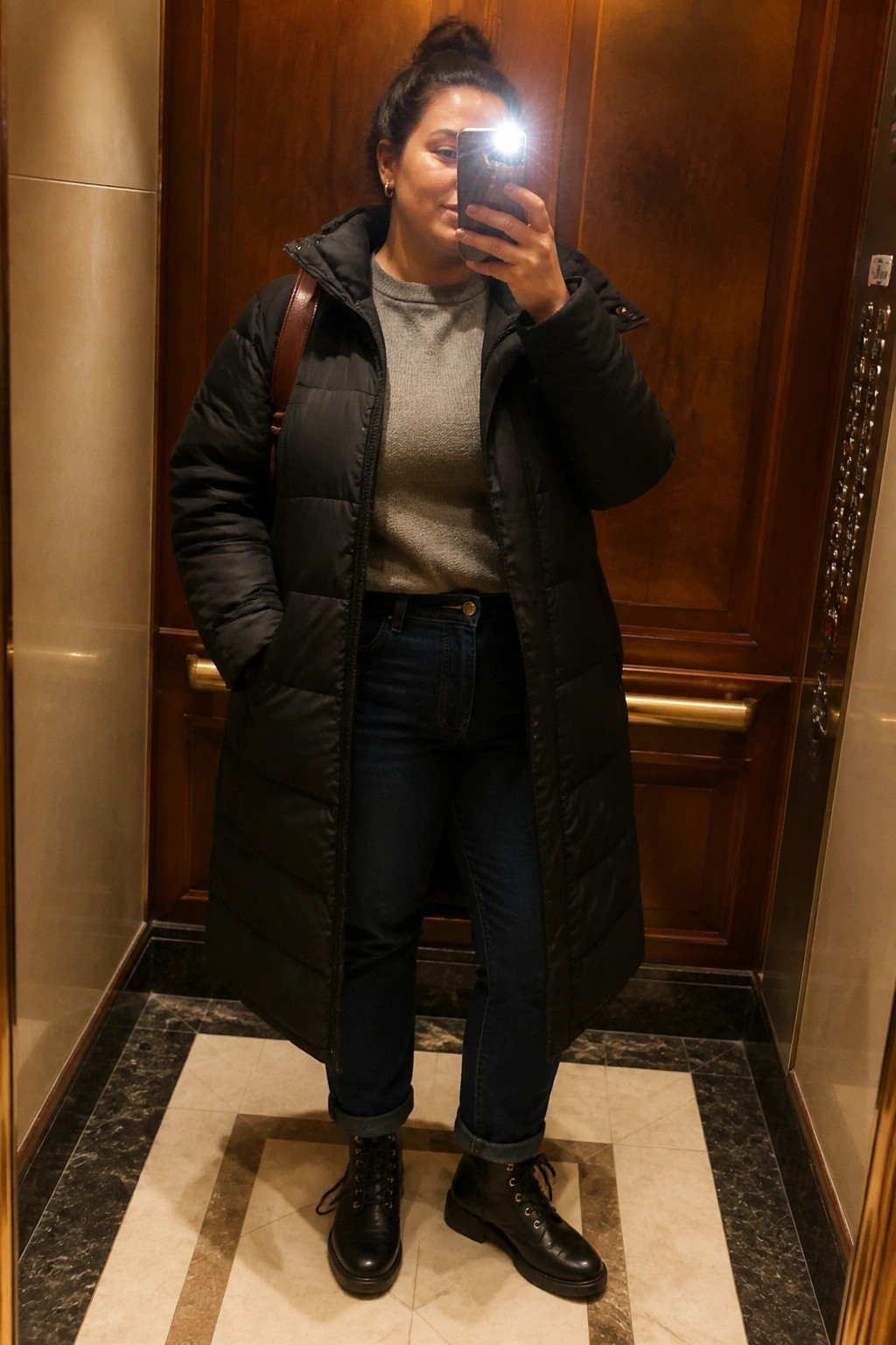 Woman in long black puffer coat taking selfie inside elevator with wooden doors