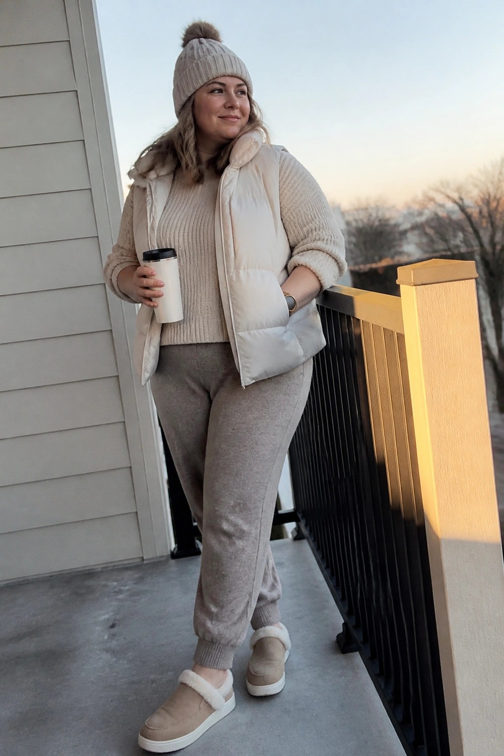 A plus size woman in beige joggers, a chunky knit sweater, and a matching puffer vest with a pom beanie and slip on sneakers holding a coffee cup.