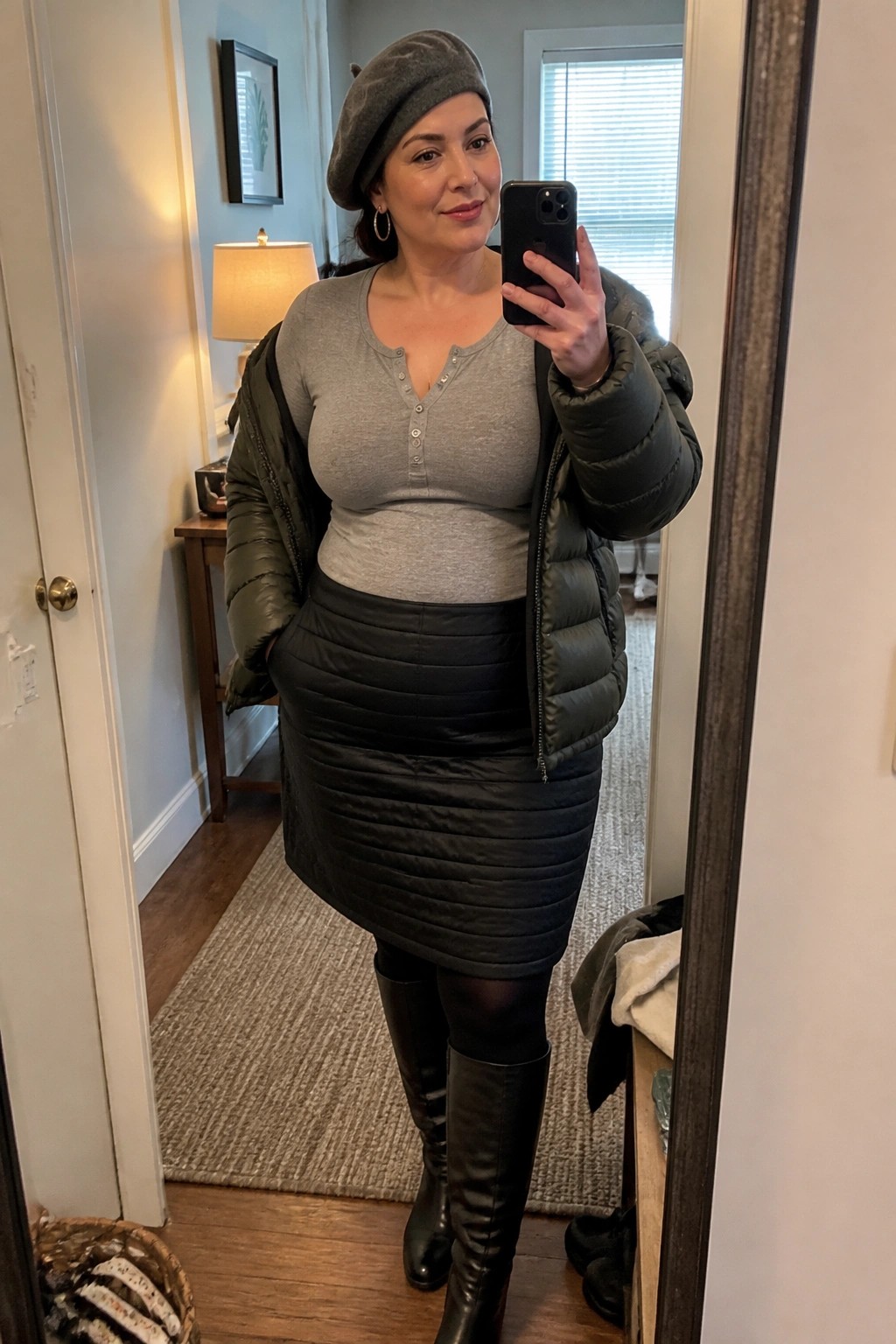 Woman wearing an open olive green puffer jacket over a gray henley top tucked into a black quilted mini skirt with black tights and knee high boots plus a gray beret while taking a mirror selfie.