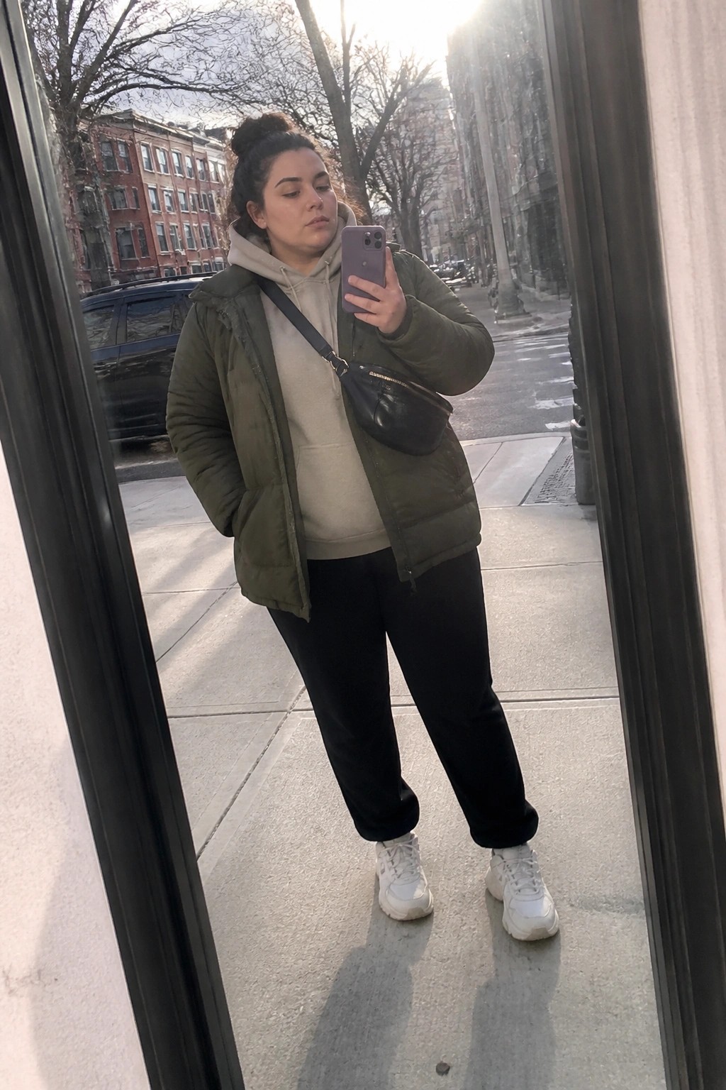 Woman in green puffer jacket and beige hoodie taking mirror selfie on city sidewalk
