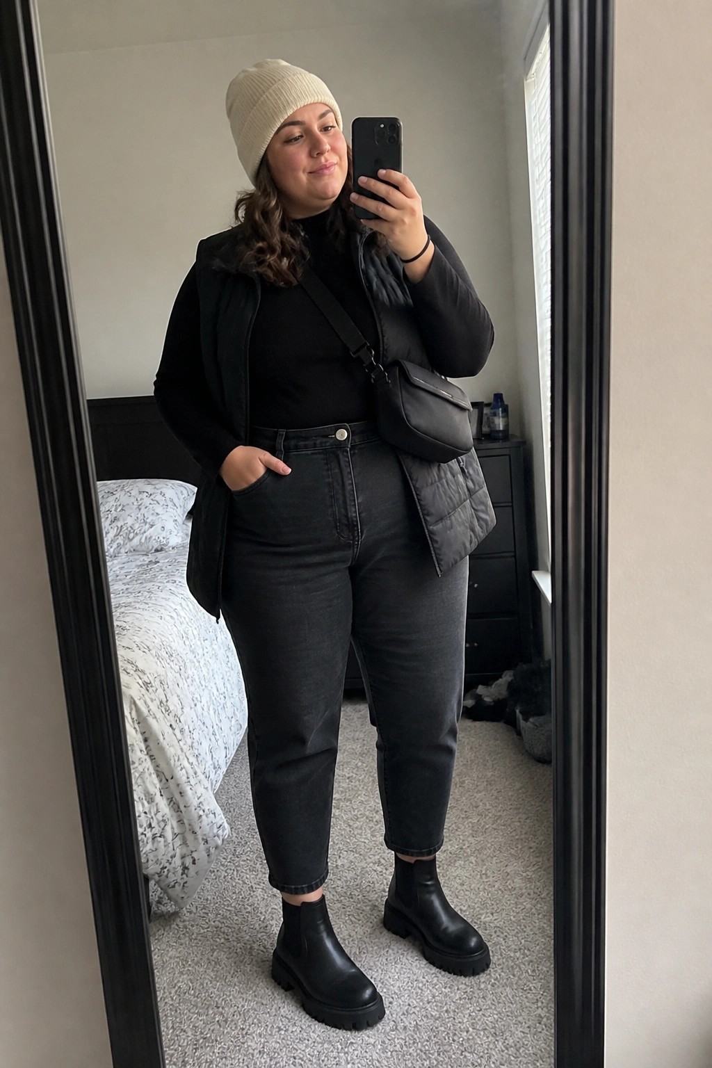 Woman in cream beanie, black vest, and jeans taking bedroom mirror selfie