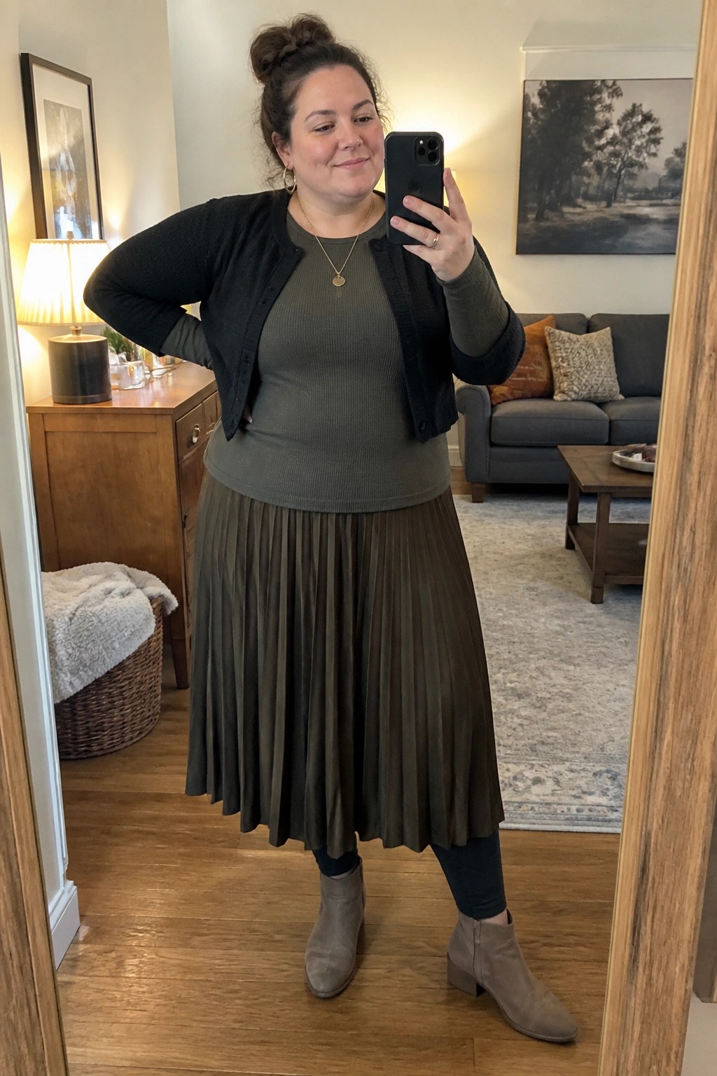 Woman in olive green pleated skirt and black cardigan taking mirror selfie in living room
