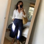 Woman in white button-up shirt tucked into navy wide-leg cropped pants with brown slide sandals and a woven straw bag, taking a mirror selfie.