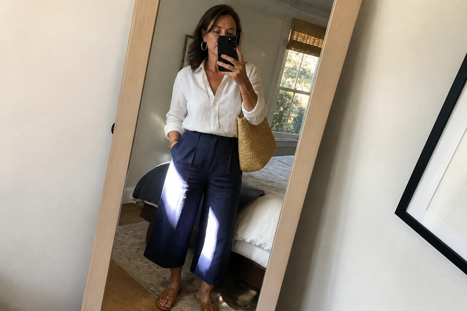 Woman in white button-up shirt tucked into navy wide-leg cropped pants with brown slide sandals and a woven straw bag, taking a mirror selfie.